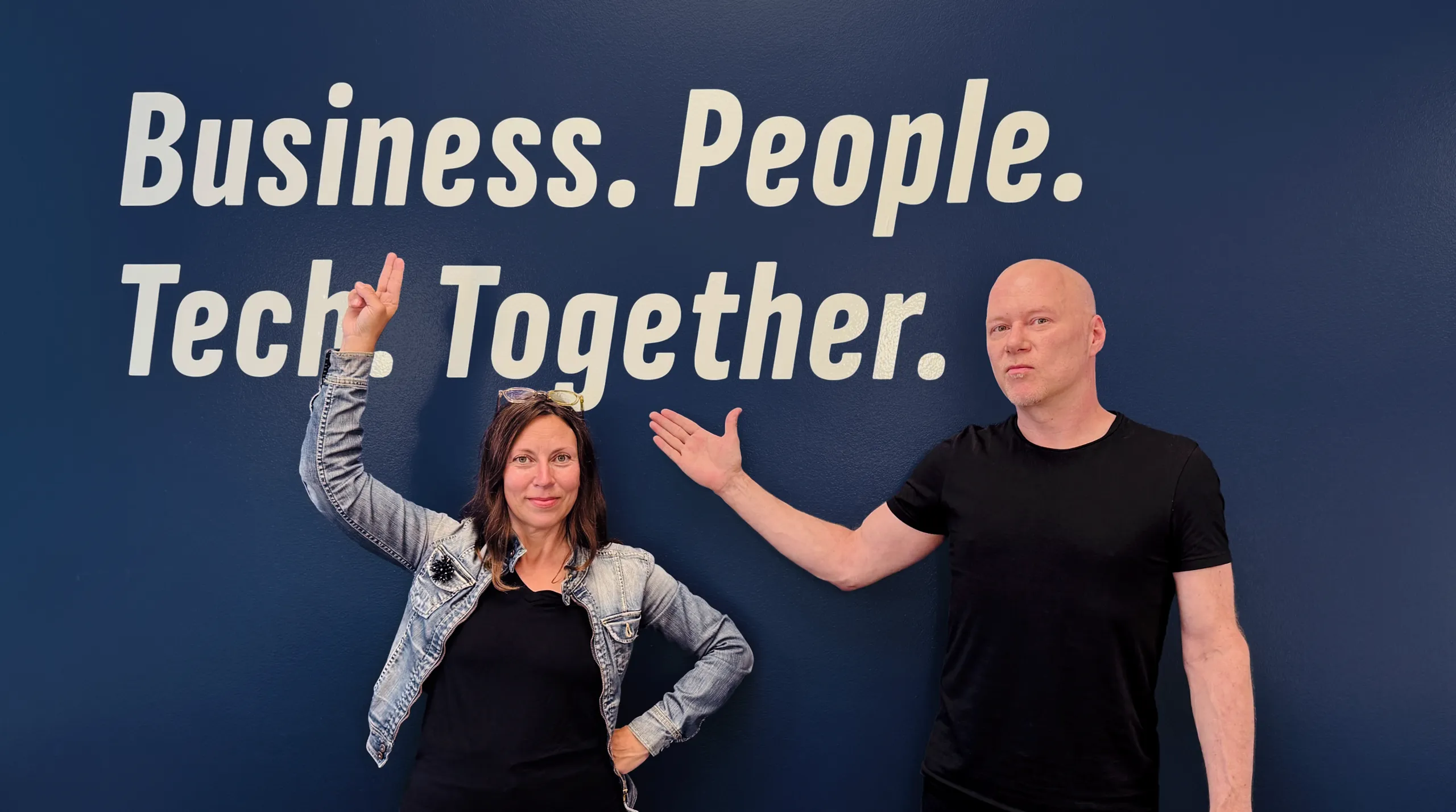 Two people posing in front of a blue wall with text reading "Business. People. Tech. Together."