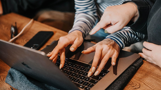 Person typing on laptop, other pointing at screen.