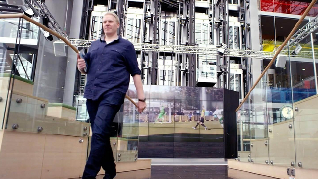  Man walking in office building with football streaming on big screen.