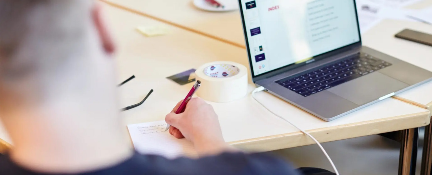 Man is writing by the table next to a open laptop