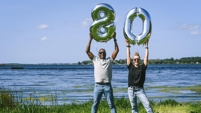Two happy Mearra people standing next to the river on a sunny day, one of the persons holding foil balloon with number eight, another person holding number zero